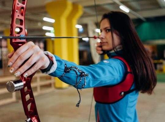 A woman in a blue jacket aims a bow and arrow with concentration during indoor archery practice.