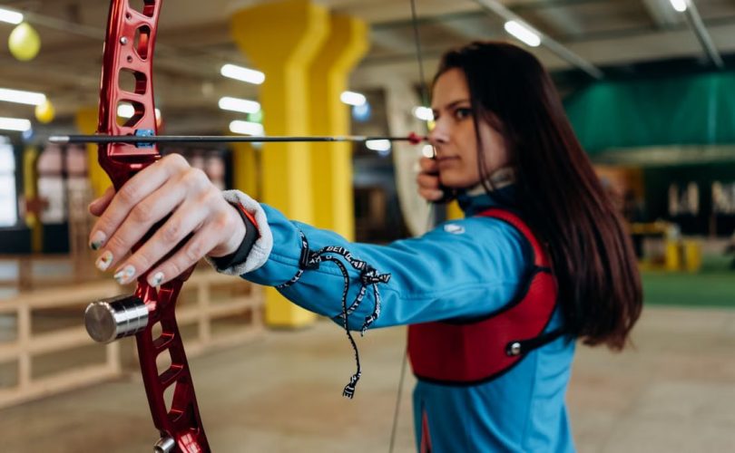 A woman in a blue jacket aims a bow and arrow with concentration during indoor archery practice.
